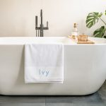 White personalized bath towel with the name "Ivy" embroidered in blue, draped over a modern bathtub.