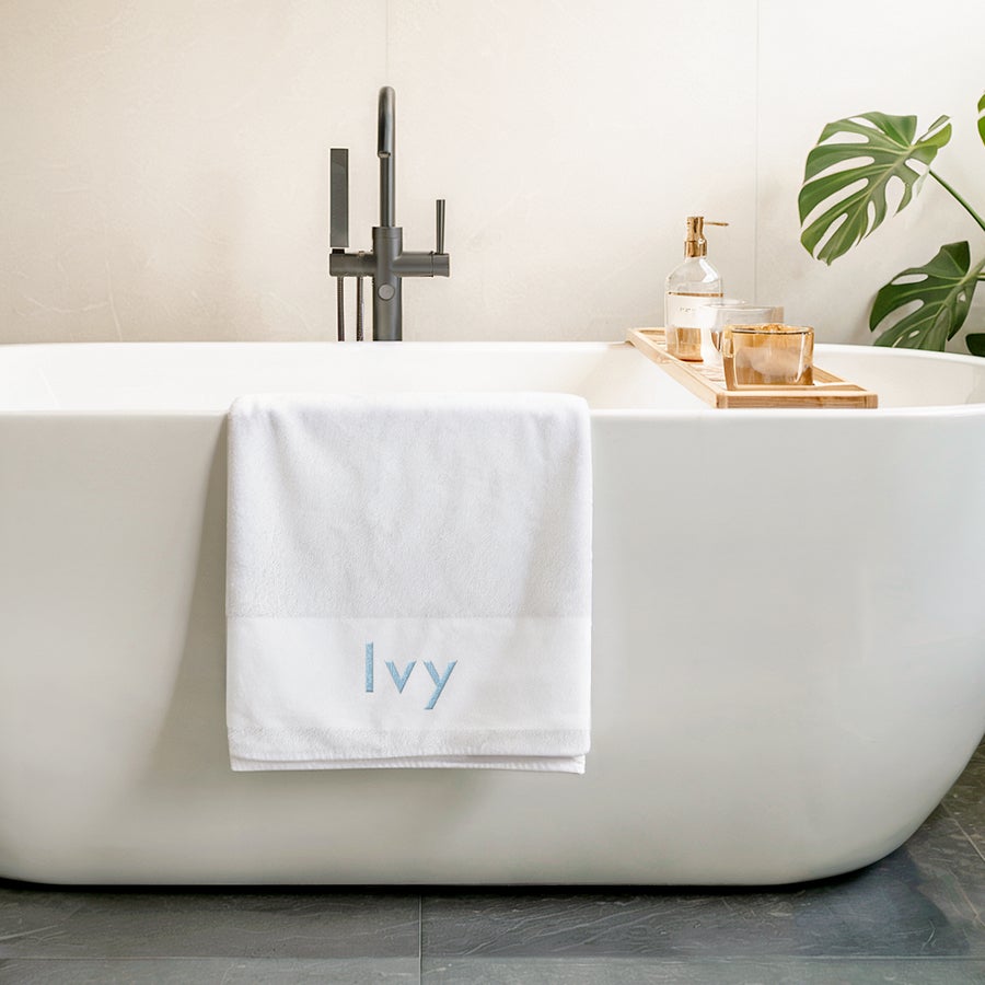White personalized bath towel with the name "Ivy" embroidered in blue, draped over a modern bathtub.