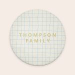 Round personalised bath mat with a cream background and blue grid pattern, printed with THOMPSON FAMILY in gold text
