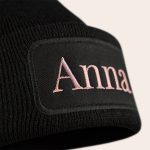 Black knitted beanie embroidered with the name Anna in pink thread on a dark fabric patch