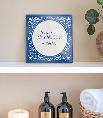 A personalized ceramic tile with a blue ornamental border and the proverb "There's no place like home - Rachel" printed in the center.