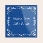 A blue personalized ceramic tile printed with "Welcome home Luke & Amy" and decorative white scrollwork