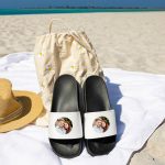 Black and white personalised sliders with a printed photo of a couple, shown on a beach towel with a hat and bag, ideal for summer.