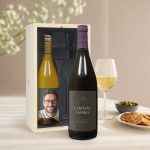 Two Salentein wine bottles in a wooden box, one with a personalized photo label and one red wine with a Chateau Harris label, with a glass of white wine and snacks.