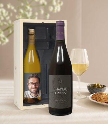 Two Salentein wine bottles in a wooden box, one with a personalized photo label and one red wine with a Chateau Harris label, with a glass of white wine and snacks.