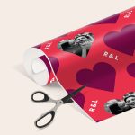 Personalised wrapping paper printed with a custom photo and initials R&L on a red heart pattern, being cut by scissors.